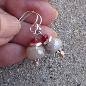May include: A pair of dangle earrings featuring gray, pearlescent beads with silver accents and small red beads. The earrings have silver hooks and are held up by a hand.
