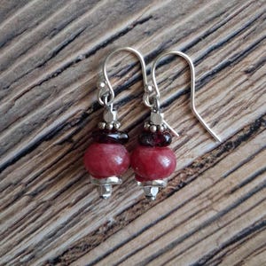 May include: A pair of dangle earrings with red and silver accents. Each earring features a large, round, red bead, a smaller dark red bead, and silver-colored metal components. The earrings hang from silver-colored ear wires.