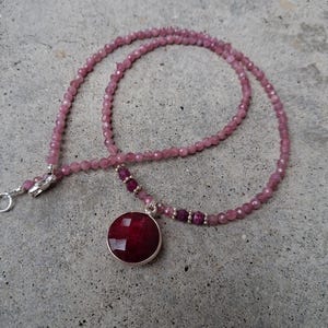 May include: A delicate necklace featuring a round, faceted, deep red pendant set in a silver-colored bezel. The necklace has a strand of small, light pink beads, with darker pink beads near the pendant. The clasp is silver-colored.