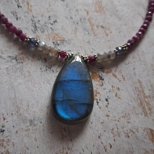 May include: A necklace featuring a large, teardrop-shaped blue labradorite pendant. The necklace has a beaded strand of small, round, dark red beads, with accents of smaller, lighter-colored beads. The pendant is the focal point.
