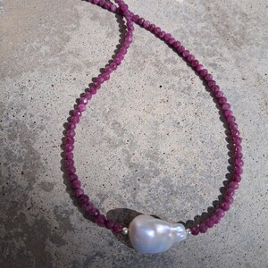 May include: A necklace featuring small, faceted, deep pink beads strung together. A large, irregular-shaped, white pearl is suspended from the necklace. The necklace is secured with a silver clasp. The jewelry is displayed on a gray surface.