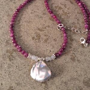 May include: A necklace featuring a strand of small, faceted, deep magenta-colored beads. The center of the necklace displays a large, irregular, iridescent white baroque pearl pendant flanked by two small, translucent, pale grey beads and silver accents.