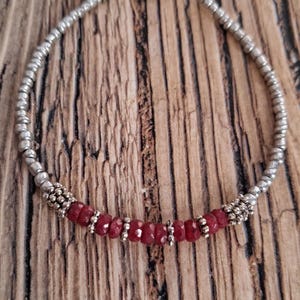 May include: Silver beaded bracelet with a section of red gemstone beads. The bracelet has a lobster clasp closure and a small red gemstone bead at the end. The bracelet is on a wooden surface.