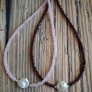 May include: Two necklaces with different colored beads and a large white pearl pendant. One necklace has light pink beads, and the other has dark red beads. The necklaces are displayed on a wooden surface.