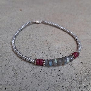 May include: Silver beaded bracelet with a mix of red and gray gemstone beads. The bracelet features a lobster clasp closure. The gemstones are a mix of ruby and labradorite. The bracelet is approximately 7 inches in length.