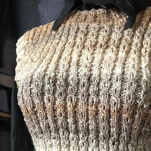 May include: A hand-knitted vest in shades of brown, beige, and cream. The sleeveless garment features a textured, open-weave design with vertical ribbing and a fitted waist. The vest is displayed on a mannequin over a black collared shirt.