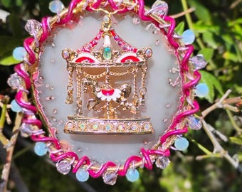 May include: A decorative carousel ornament with a gold-colored base and a red and white roof. The carousel features miniature horses and is surrounded by a pink and gold frame adorned with crystals and blue beads. The ornament hangs from a pink cord.