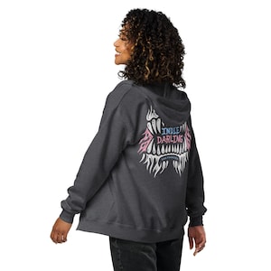 Könnte beinhalten: Dunkelgrauer Kapuzenpullover mit Reißverschluss und Kapuze. Auf der Rückseite befindet sich ein grafisches Design mit den Worten "Indie Darling" in Rosa und Blau sowie "San Francisco" in Weiß. Das Design hat eine stilisierte, zahnartige Form.