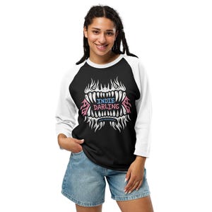 May include: Black and white raglan shirt with the text "INDIE DARLING" in a stylized design. The design includes pink and blue accents and the words "SAN FRANCISCO". The shirt is paired with denim shorts.