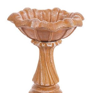 May include: A brown concrete bird bath with a scalloped edge and a pedestal base. The bird bath has a rustic, weathered look.