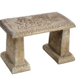 May include: A decorative stone bench with an intricate carved design. The bench is made of a light beige stone and has a rectangular seat and two legs on each side.