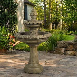 May include: A stone garden fountain with a three-tiered design. The fountain has a pedestal base and is surrounded by greenery, including potted plants and a stone wall. The fountain is made of a light-colored stone.