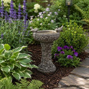 May include: A gray stone bird bath with two small bird figurines on the rim. The bird bath is in a garden setting, surrounded by green plants, purple flowers, and a stone pathway. A black solar lamp is in the background.