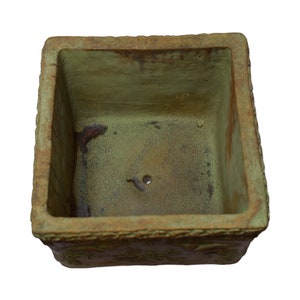 May include: A square, weathered planter with a textured, olive-green interior and a decorative border. The planter has a drainage hole and is suitable for plants. The exterior has a rustic, aged appearance. The planter is suitable for plants.