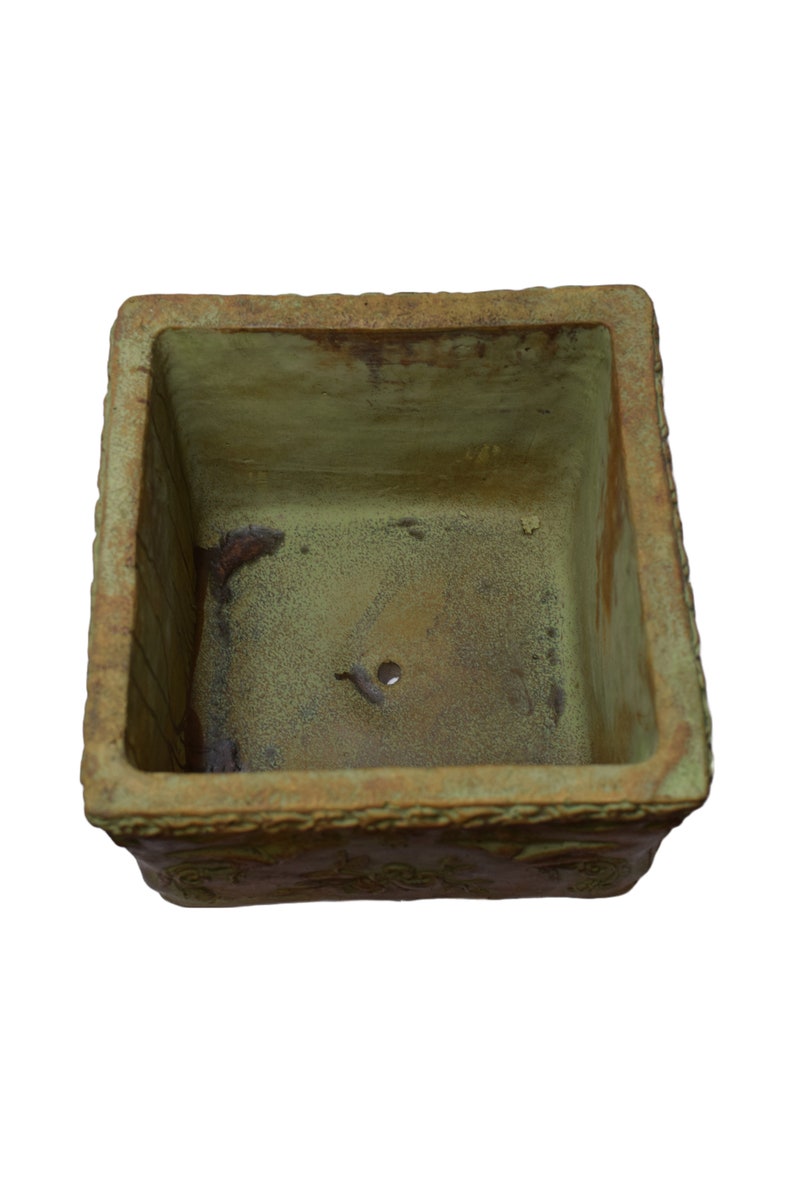 May include: A square, weathered planter with a textured, olive-green interior and a decorative border. The planter has a drainage hole and is suitable for plants. The exterior has a rustic, aged appearance. The planter is suitable for plants.