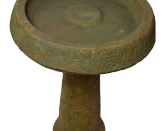 May include: A green concrete bird bath with a wide, shallow bowl and a tall, cylindrical pedestal. The bowl has a textured surface and the pedestal is decorated with a floral pattern.
