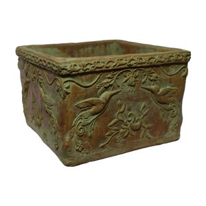 May include: Square planter with a distressed finish, featuring a hummingbird and floral relief pattern. The planter has a brown base with green highlights, and a decorative border. Suitable for gardens or interior spaces.