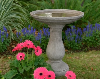 May include: A gray stone bird bath filled with water stands in a garden. Bright pink gerbera daisies bloom in the foreground, with blue flowers and green foliage surrounding the bird bath.