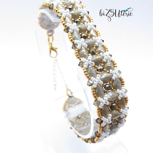 Op de afbeelding: Een grijze en gouden kralen armband met een witte achtergrond. De armband is gemaakt met kleine, ronde kralen en heeft een geometrisch patroon. De armband staat op een witte displaystandaard.