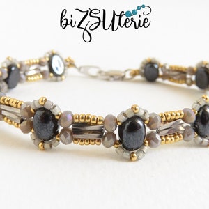 May include: A beaded bracelet featuring oval black stones, gray beads, and gold accents. The bracelet has a silver clasp and the brand name "bizZutlerie" is in the upper left corner.