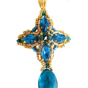 May include: A gold pendant with a cross-shaped design, incorporating large, faceted teal-blue gemstones and smaller green and blue beads. A teardrop-shaped teal-blue gemstone hangs at the bottom. The text "zohara" and "beading pattern by b'zSuterie design" are visible.