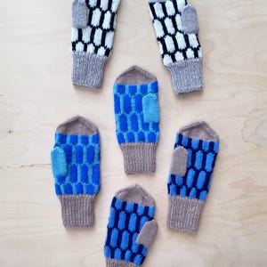 May include: Six knitted mittens in shades of blue, grey, and white are arranged on a light wood surface. The mittens feature geometric patterns, with some displaying a honeycomb design and others a rectangular pattern. The cuffs and thumbs are a solid grey colour.
