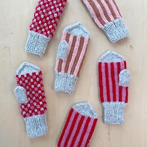 May include: Six pairs of knitted mittens in various patterns. The mittens are grey, pink, and red with white accents. The mittens are arranged in a semi-circle on a light wood surface.