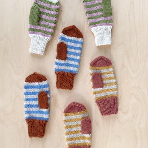 Könnte beinhalten: Fünf gestrickte Fäustlinge in einer Anordnung. Zwei Fäustlinge haben weiße, grüne und violette Streifen mit weißen Bündchen. Zwei Fäustlinge haben blaue und weiße Streifen mit braunen Bündchen. Ein Fäustling hat gelbe und weiße Streifen mit mauvefarbenem Bündchen.