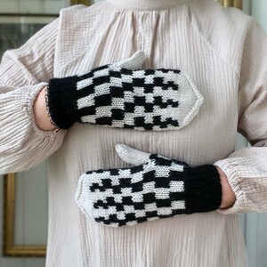 May include: A pair of black and white checked knitted mittens. The mittens are held in front of a person wearing a light beige shirt.