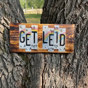 May include: A wooden sign with the words "GET LE1D" spelled out using license plates. The sign is attached to a tree trunk.