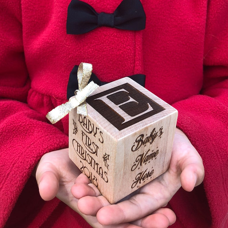 May include: A light-colored wooden cube ornament featuring a dark engraved letter E on the top surface. The side panels are engraved with the text Baby's First Christmas 2018 and Baby's Name Here. A small gold ribbon is attached to the top corner.