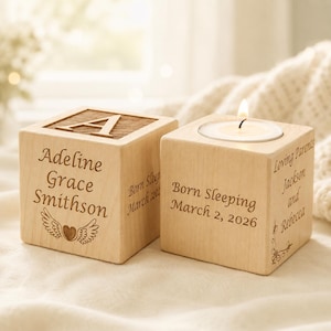 May include: Two light-colored wooden blocks. One block features the letter A on top and the name Adeline Grace Smithson with a heart and wings graphic. The other block is a candle holder with a lit tea light, inscribed with Born Sleeping March 2, 2026, and Loving Parents Jackson and Rebecca.