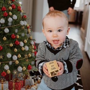 May include: A small wooden block with the engraved name Aiden Lee Miller and a large letter A on the front. The block is light brown with dark lettering and is held in front of a decorated Christmas tree with red, gold, and silver ornaments.