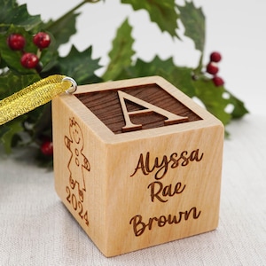 May include: A light-colored wooden block ornament featuring a dark wood top with the letter A engraved. One side displays the name Alyssa Rae Brown, while another side shows an engraved gingerbread person and the year 2024. A gold ribbon is attached.