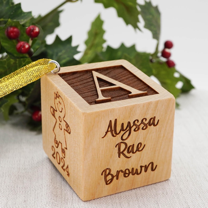 May include: A light-colored wooden block ornament featuring a dark wood top with the letter A engraved. One side displays the name Alyssa Rae Brown, while another side shows an engraved gingerbread person and the year 2024. A gold ribbon is attached.