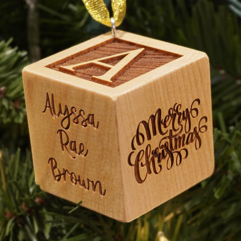 May include: A light-colored wooden cube ornament featuring a gold ribbon loop at the top. The top face displays a large letter A. One side is engraved with the name Alyssa Rae Brown, and another side features the text Merry Christmas in script.