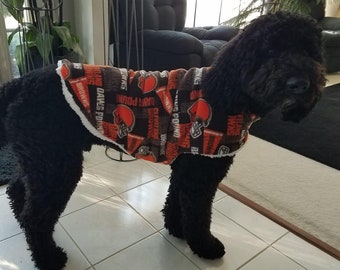 May include: A black dog wearing a dog coat. The coat is brown and orange with the words "BROWNS" and "DAWGS POUND" printed on it. The coat has a white trim.