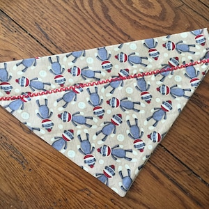 May include: A tan triangular dog bandana with a red stitched edge. The bandana is covered in a pattern of gray sock monkeys with red hats and white buttons.