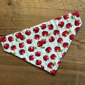 May include: A white fabric dog bandana with a red apple print on a woodgrain background. The bandana has a red stitched edge.