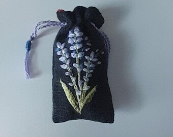 May include: A small black fabric pouch with a lavender flower embroidered on the front. The pouch has a drawstring closure and a purple ribbon.