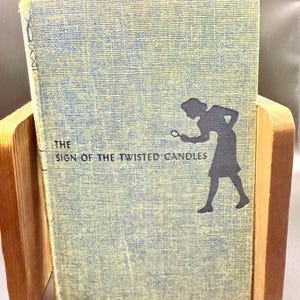 May include: A vintage book titled "The Sign of the Twisted Candles" is displayed in a wooden book holder. The book cover is light blue with a silhouette of a person holding a magnifying glass. The book has a textured appearance.