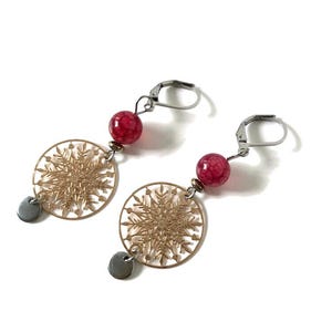 May include: Dangle earrings with a gold snowflake design, red beads, and small grey discs. The earrings have a lever-back closure. The earrings are set against a white background. Each earring has a gold snowflake design.