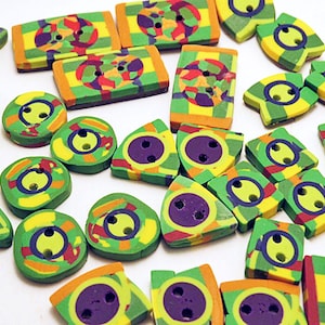 Op de afbeelding: Een verzameling diverse knopen in verschillende geometrische vormen, waaronder cirkels, rechthoeken en driehoeken. Elke knoop heeft een levendig, meerkleurig patroon van groen, geel, oranje en paars, met twee gaatjes om vast te naaien.