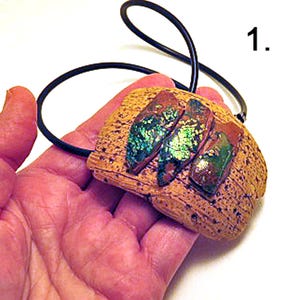 Pu&ograve; includere: Un ciondolo con una base testurizzata color beige, simile all'argilla, con tre inserti rettangolari iridescenti dai riflessi metallici verdi e blu. Il ciondolo &egrave; fissato a un semplice cordino nero. Il numero 1 &egrave; stampato nell'angolo in alto a destra.
