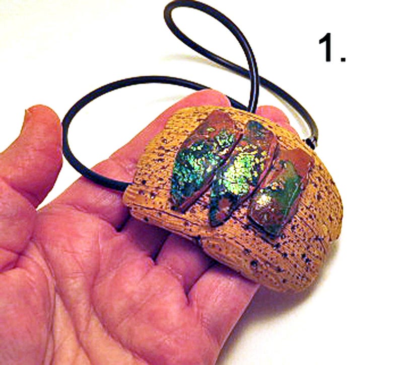 Pu&ograve; includere: Un ciondolo con una base testurizzata color beige, simile all'argilla, con tre inserti rettangolari iridescenti dai riflessi metallici verdi e blu. Il ciondolo &egrave; fissato a un semplice cordino nero. Il numero 1 &egrave; stampato nell'angolo in alto a destra.