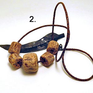 Pu&ograve; includere: Una collana con un sottile cordino in pelle intrecciata marrone, infilato con cinque perline di legno cilindriche, di colore marrone chiaro e testurizzate. Dietro le perline si trova un ciondolo metallico scuro a forma di foglia.