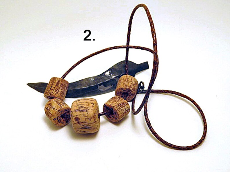 Pu&ograve; includere: Una collana con un sottile cordino in pelle intrecciata marrone, infilato con cinque perline di legno cilindriche, di colore marrone chiaro e testurizzate. Dietro le perline si trova un ciondolo metallico scuro a forma di foglia.