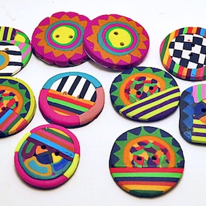 Puede incluir: Una colección de diez botones redondos con patrones geométricos, rayas y diseños de rayos de sol en colores vibrantes. Cada botón tiene dos agujeros centrales y muestra una variedad de colores brillantes que incluyen rosa, amarillo, verde, azul y negro.