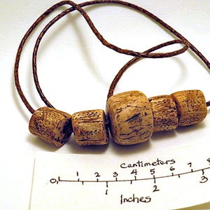 Pu&ograve; includere: Una collana con cinque perline cilindriche testurizzate color marrone chiaro su un cordino intrecciato marrone scuro. Sotto le perline &egrave; posizionato un righello di carta con scala in centimetri e pollici, con la perla pi&ugrave; grande di 1,3 cm.