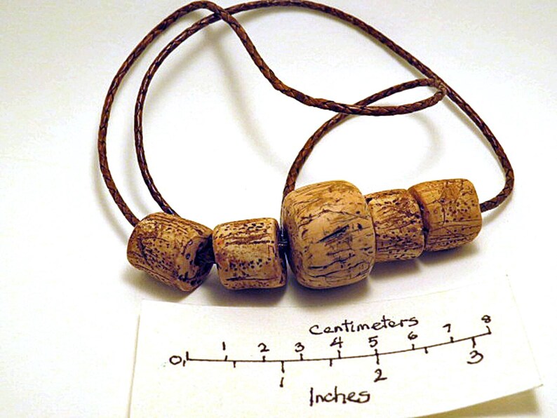 Pu&ograve; includere: Una collana con cinque perline cilindriche testurizzate color marrone chiaro su un cordino intrecciato marrone scuro. Sotto le perline &egrave; posizionato un righello di carta con scala in centimetri e pollici, con la perla pi&ugrave; grande di 1,3 cm.